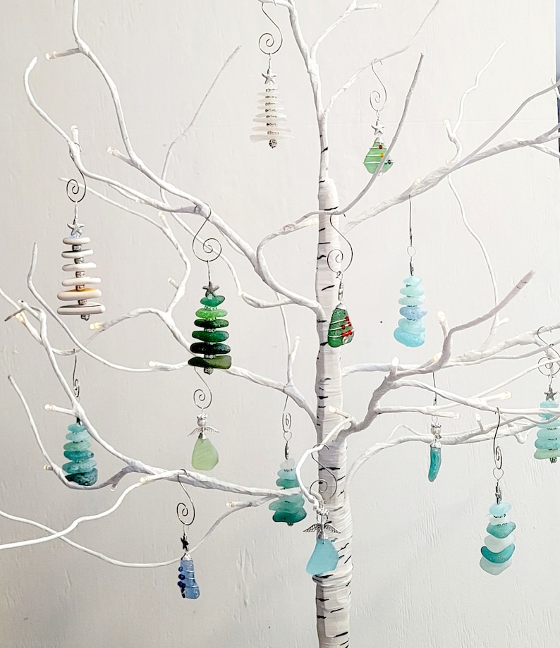 May include: A white twig tree with 12 glass ornaments shaped like Christmas trees. The ornaments are decorated with green, blue, and white glass pieces.