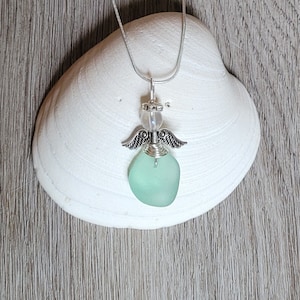 Pode incluir: Um colar com um pingente de anjo de prata e um charme de vidro do mar verde claro. O pingente está pendurado em uma corrente de prata.