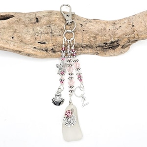 May include: A beach-themed keychain with a silver clasp and charms. The charms include a starfish, seashell, and a piece of sea glass. Pink beads and a pink gem accent the design. The keychain is displayed on a piece of driftwood.