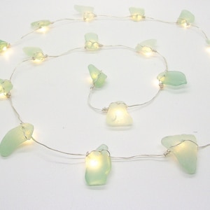 May include: A string of fairy lights featuring sea glass pieces in shades of green and white. Each piece is illuminated by a small, warm white LED light. The lights are strung on a thin, silver wire.
