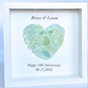 May include: A square white shadow box frame containing a heart-shaped arrangement of pale green and light blue sea glass pieces. The text Brian & Laura is printed above the heart, and Happy 10th Anniversary 06.25.2016 is printed below the heart.