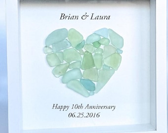 May include: A square white shadow box frame containing a heart-shaped arrangement of pale green and light blue sea glass pieces. The text Brian & Laura is printed above the heart, and Happy 10th Anniversary 06.25.2016 is printed below the heart.