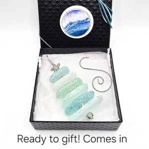 May include: A sea glass Christmas tree ornament in a black gift box with a white satin lining. The ornament is made of five pieces of sea glass in shades of blue and green, with a silver starfish on top and a silver seashell on the bottom. The ornament comes with a silver ornament hook.