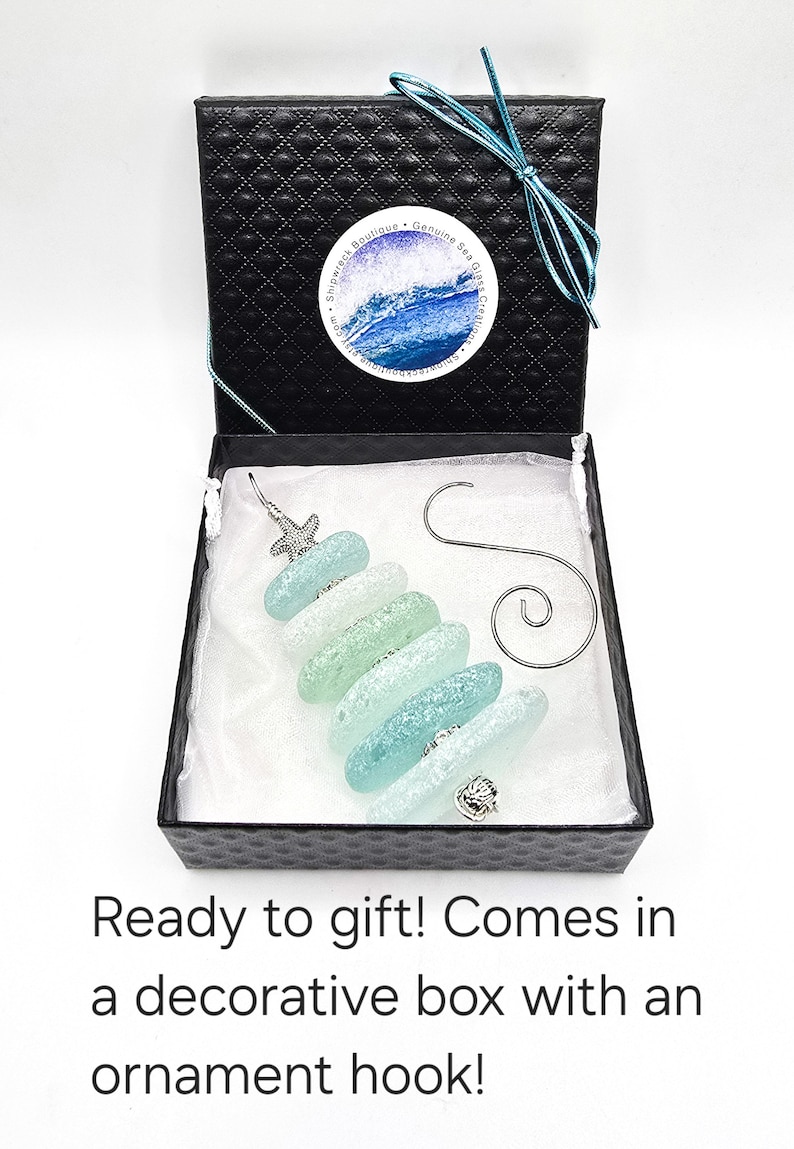 May include: A sea glass Christmas tree ornament in a black gift box with a white satin lining. The ornament is made of five pieces of sea glass in shades of blue and green, with a silver starfish on top and a silver seashell on the bottom. The ornament comes with a silver ornament hook.
