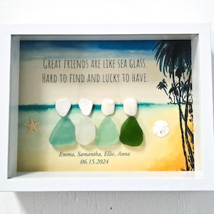 Puede incluir: Obra de arte enmarcada con figuras de vidrio marino en una escena de playa. El texto dice: "Los buenos amigos son como el vidrio marino. Difíciles de encontrar y afortunados de tener." Se incluyen nombres y una fecha. El marco es blanco.