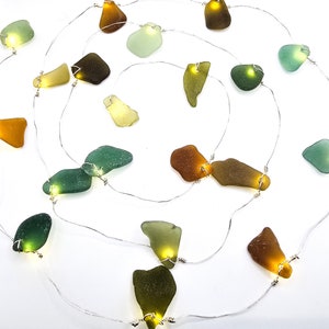 May include: A decorative string of fairy lights with pieces of sea glass in shades of green, brown, and yellow. Each piece has a small LED light, producing a warm glow. The lights are strung on a thin silver wire, suitable for indoor decoration.