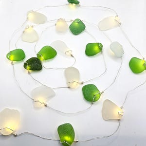 May include: A string of fairy lights featuring sea glass in shades of green and white. Each piece of sea glass is illuminated by a small LED light. The lights are strung on a thin, silver wire, creating a decorative and whimsical effect.