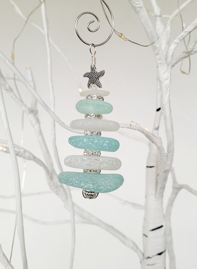 May include: A teal and white sea glass Christmas tree bauble with a silver starfish and silver beads. The bauble is hanging from a silver wire hanger.