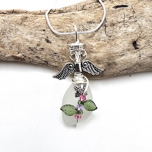 Pode incluir: Um colar com pingente de anjo prateado feito à mão. O pingente apresenta uma cabeça de cristal transparente, asas detalhadas e um corpo de vidro do mar. Enfeitado com contas rosa e roxas e folhas verdes, ele pende de uma corrente prateada.