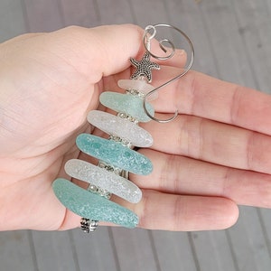 May include: A sea glass Christmas tree bauble with a silver starfish on top. The bauble is made of five pieces of sea glass in shades of blue and white, with silver beads between each piece.