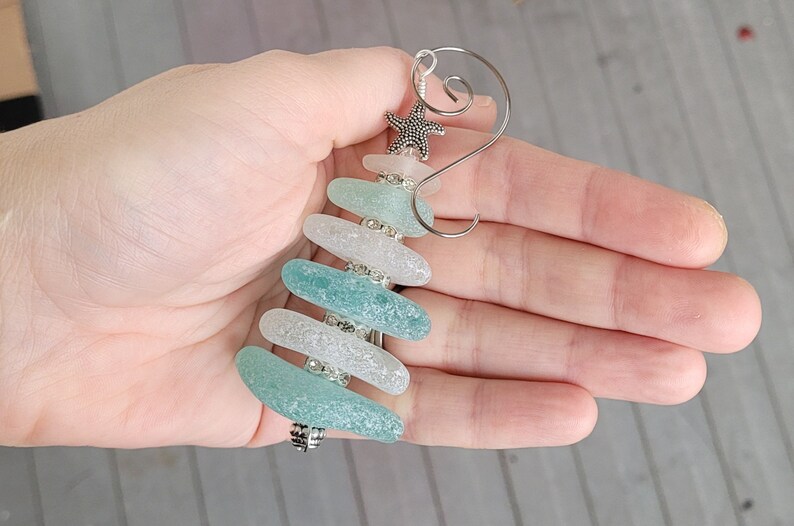 May include: A sea glass Christmas tree bauble with a silver starfish on top. The bauble is made of five pieces of sea glass in shades of blue and white, with silver beads between each piece.