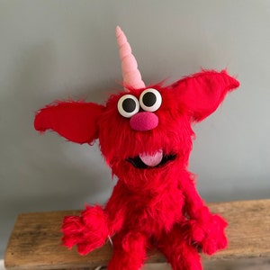 May include: A red furry puppet with a pink unicorn horn, large black eyes, and a pink tongue sticking out. The puppet is sitting on a wooden bench.