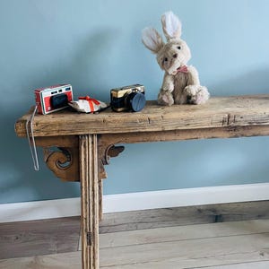 May include: A light brown bunny rabbit wearing a pink bow tie sits on a wooden bench with two vintage cameras and a red and white gift bag.