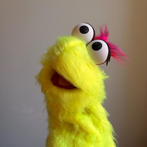 Puede incluir: Una marioneta de mano de color amarillo neón brillante con pelo largo sintético, dos grandes ojos esféricos blancos con pupilas negras, un mechón de pelo rosa vibrante en la parte superior de la cabeza y una boca abierta de color rojo oscuro.
