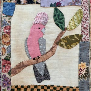 May include: A pink and gray bird with a pink crest perched on a brown branch with green leaves. The bird is surrounded by a patchwork of fabric squares in various colors and patterns.