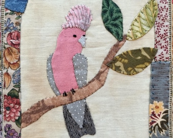 May include: A pink and grey bird with a pink crest perched on a brown branch with green leaves. The bird is surrounded by a patchwork of fabric squares in various colours and patterns.