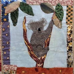 May include: A gray koala bear is climbing a brown tree with green leaves. The koala is made of fabric and is stitched with brown thread. The tree is made of fabric and is stitched with yellow thread. The background is a light blue fabric.