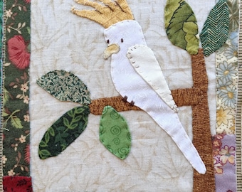 May include: A white cockatoo with a yellow crest perched on a brown branch with green leaves. The bird is sitting on a white fabric background.
