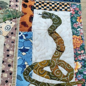 May include: A patchwork quilt with a green snake design in the center. The quilt is made of various fabrics with floral and geometric patterns in shades of blue, red, green, brown, and yellow.