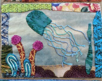 May include: A blue and green jellyfish with long tentacles is embroidered on a light blue fabric background. The jellyfish is surrounded by three pink and brown coral-like shapes.