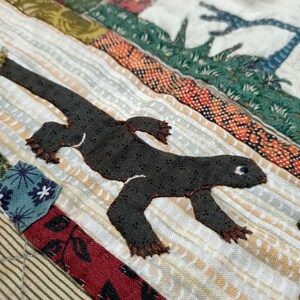 May include: A close-up of a brown and black lizard applique on a white fabric background. The lizard is stitched onto a patchwork quilt with colorful fabric squares.