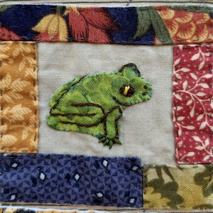 May include: A green frog embroidered on a white fabric background. The frog is surrounded by a patchwork of different fabrics in various colours and patterns.