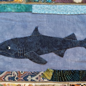 May include: A blue fabric shark appliqué on a light blue background surrounded by a colorful patchwork border.