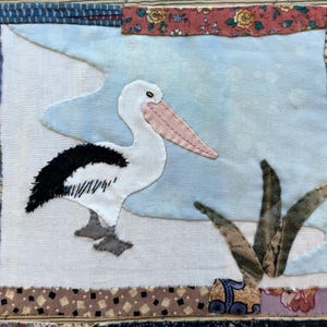 May include: A white pelican with black feathers on its back is stitched onto a white fabric background. The pelican is standing on a light blue fabric that looks like water. There are green fabric leaves in the background.