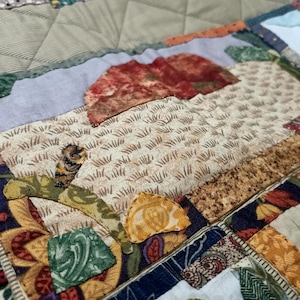 May include: A colourful patchwork quilt featuring a variety of fabrics and patterns, including a brown and white fabric with a grass-like design, a red and brown fabric with a mushroom-like design, and a blue and white fabric with a floral design.