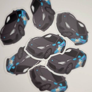 Puede incluir: Una colección de pegatinas negras para coches con un diseño de chica de anime de dibujos animados. Las pegatinas presentan un coche gris oscuro con detalles en azul y turquesa, y una chica de anime sonriente con pelo azul. Las pegatinas están dispuestas sobre una superficie blanca.