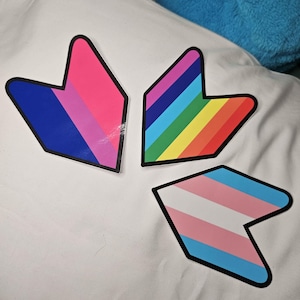 May include: Three arrow-shaped stickers with different pride flag designs. The first sticker is blue, pink, and white. The second sticker is rainbow. The third sticker is light blue, pink, and dark blue.