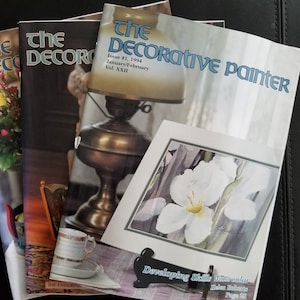May include: Three copies of The Decorative Painter magazine, a publication for artists. The front cover of the top magazine features a painting of white flowers. The text on the cover reads "The Decorative Painter", "Issue #1, 1994", "January/February", "Vol. XXII", and "Developing Skills Watercolor".