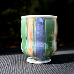 May include: A ceramic cup with a white rim and base. The cup features vertical stripes of green, orange, and blue with gold speckles. The cup is approximately 4 inches tall.