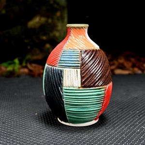 Puede incluir: Un jarrón de cerámica colorido con un diseño de patchwork. El jarrón presenta una variedad de colores que incluyen rojo, naranja, azul, verde, marrón y negro. El jarrón tiene una superficie texturizada y una pequeña abertura en la parte superior. Artículo decorativo.
