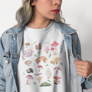 Peut inclure: Sweat-shirt blanc avec un imprimé de champignons colorés. Les champignons sont de formes et de tailles variées, et certains ont des taches rouges, roses, brunes et blanches. Le sweat-shirt a un col rond et des manches longues.