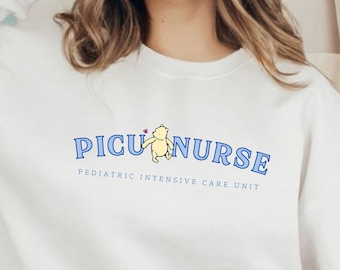 Puede incluir: Sudadera blanca con texto azul que dice "PICU NURSE PEDIATRIC INTENSIVE CARE UNIT" con un pequeño oso de peluche amarillo sosteniendo una flor azul.