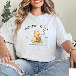 May include: A light grey t-shirt with the text "Auntie to Bee Est. 2024 A little honey is on the way" and a cartoon image of Winnie the Pooh holding a pot of honey.