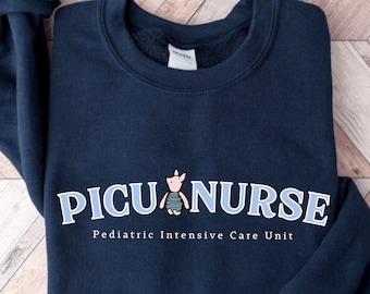 Puede incluir: Una sudadera azul marino con el texto "PICU NURSE" en letras azul claro y un pequeño cerdo de dibujos animados con una camisa a rayas. El texto "Pediatric Intensive Care Unit" está impreso debajo del texto principal.