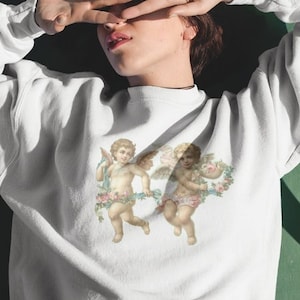 May include: White sweatshirt with a graphic print of two cherubs with wings and floral accents.