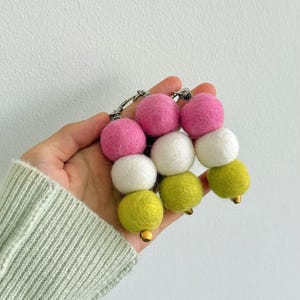 May include: A colorful keychain featuring felt balls in pink, white, and green. The felt balls are strung together with a silver chain and small gold-colored bells at the bottom. The keychain is a decorative accessory.