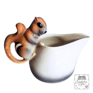 May include: A white ceramic creamer with a brown and gray squirrel handle. The squirrel is perched on the side of the creamer, with its tail forming the handle. The creamer has a slightly flared spout and a dark gray base.
