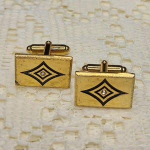 May include: A pair of vintage gold-toned cufflinks. Each cufflink features a rectangular shape with a black diamond design and a small, clear gem in the center. The cufflinks have a classic, elegant style.