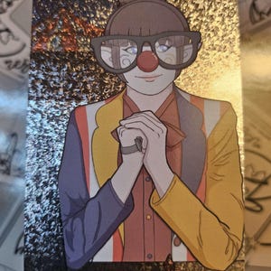 Peut inclure: Une carte &agrave; collectionner holographique rectangulaire illustrant un personnage avec un nez de clown rouge, des lunettes &agrave; monture fonc&eacute;e et les cheveux en chignon. Le personnage porte une veste multicolore aux panneaux violets, jaunes et orange.
