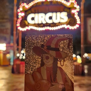 Peut inclure: Une carte &agrave; collectionner rectangulaire avec une finition &agrave; paillettes dor&eacute;es illustrant un clown portant un chapeau ray&eacute; et tenant un animal en ballon orange. La carte est tenue devant un fond flou avec une enseigne lumineuse CIRCUS.