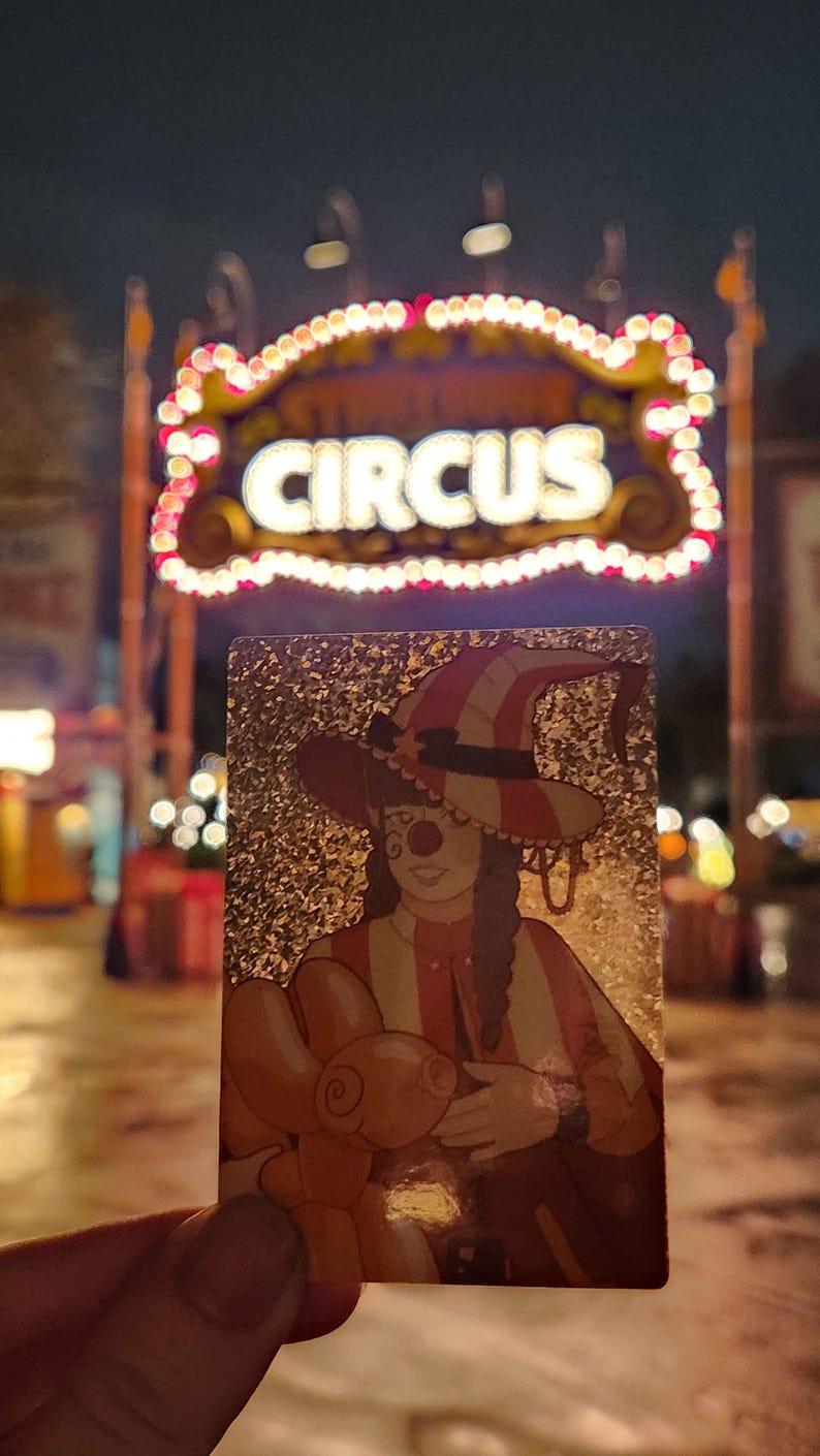 Peut inclure: Une carte &agrave; collectionner rectangulaire avec une finition &agrave; paillettes dor&eacute;es illustrant un clown portant un chapeau ray&eacute; et tenant un animal en ballon orange. La carte est tenue devant un fond flou avec une enseigne lumineuse CIRCUS.