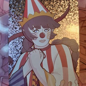 Peut inclure: Un autocollant holographique rectangulaire repr&eacute;sentant un personnage d&eacute;guis&eacute; en clown avec un chapeau conique ray&eacute; rouge et blanc, un nez rouge et des cheveux tress&eacute;s. Le personnage porte une tenue ray&eacute;e aux accents jaunes et rouges.
