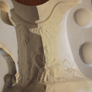 May include: A light beige ceramic mold with a tree-like structure and relief carvings of figures and foliage. The mold has a curved top and several circular indentations, suggesting a decorative or artistic purpose.