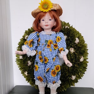 May include: A porcelain doll wearing a blue and white gingham dress with sunflowers, a straw hat with a sunflower accent, and white lace socks. The doll is standing in front of a green wreath.