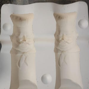 May include: A white ceramic mold featuring two identical recessed impressions of a chef wearing a tall hat and a neckerchief. The mold has a smooth, matte finish with defined facial features, including a mustache, visible in both cavities.