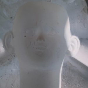 May include: A white, blank doll head mold with visible facial features, including eyes, a nose, and a mouth. The mold is set within a larger, textured form, suggesting a crafting or artistic context.